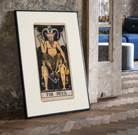 The Devil tarot card figure print in a black frame leaning beside a vintage door on parquet