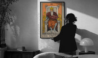 The Emperor print in a slim black frame being hung on a sunlit wall in a monochrome room
