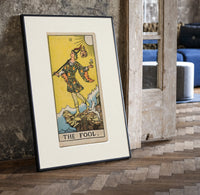 The Fool tarot card figure print in a black frame leaning beside a vintage door on parquet