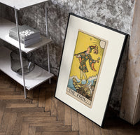 The Fool tarot card figure poster in a black frame leaning beside a shelf in a raw-plaster loft