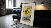 The Fool tarot card figure print in a slim black frame leaning on a dark sofa in a concrete print studio