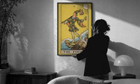 The Fool print in a slim black frame being hung on a sunlit wall in a monochrome room