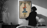 The Hanged Man print in a slim black frame being hung on a sunlit wall in a monochrome room