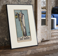 Cloaked hermit holding lantern and staff print in a black frame leaning beside a vintage door on parquet