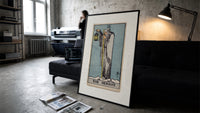 Cloaked hermit holding lantern and staff print in a slim black frame leaning on a dark sofa in a concrete print studio