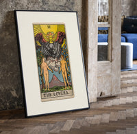 The Lovers tarot card print in a black frame leaning beside a vintage door on parquet