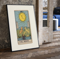 The Moon tarot card print in a black frame leaning beside a vintage door on parquet