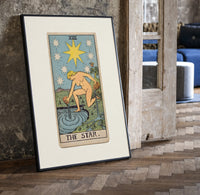 The Star tarot card figure print in a black frame leaning beside a vintage door on parquet