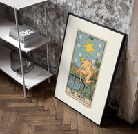 The Star tarot card figure poster in a black frame leaning beside a shelf in a raw-plaster loft