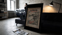 Santa in sleigh and figure riding goose print in a slim black frame leaning on a dark sofa in a concrete print studio