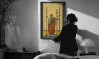Three of Wands print in a slim black frame being hung on a sunlit wall in a monochrome room