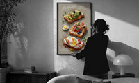 Three Toasts with Egg, Avocado and Fig print in a slim black frame being hung on a sunlit wall in a monochrome room