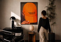 Trusting God Orange Cap print in a black frame on a sunlit plaster wall, a woman looking on beside a mushroom lamp