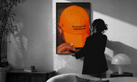 Trusting God Orange Cap print in a slim black frame being hung on a sunlit wall in a monochrome room