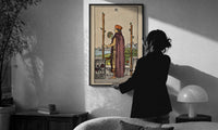 Two of Wands print in a slim black frame being hung on a sunlit wall in a monochrome room