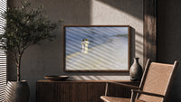 Two women walking on beach print in a wood frame above a fluted sideboard beside an olive tree