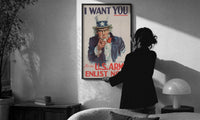 Uncle Sam I Want You print in a slim black frame being hung on a sunlit wall in a monochrome room