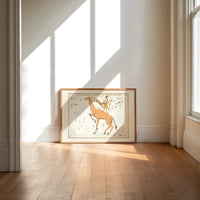 Camelopardalis constellation giraffe with figures print in an oak frame on a sunlit wood floor beneath tall window light