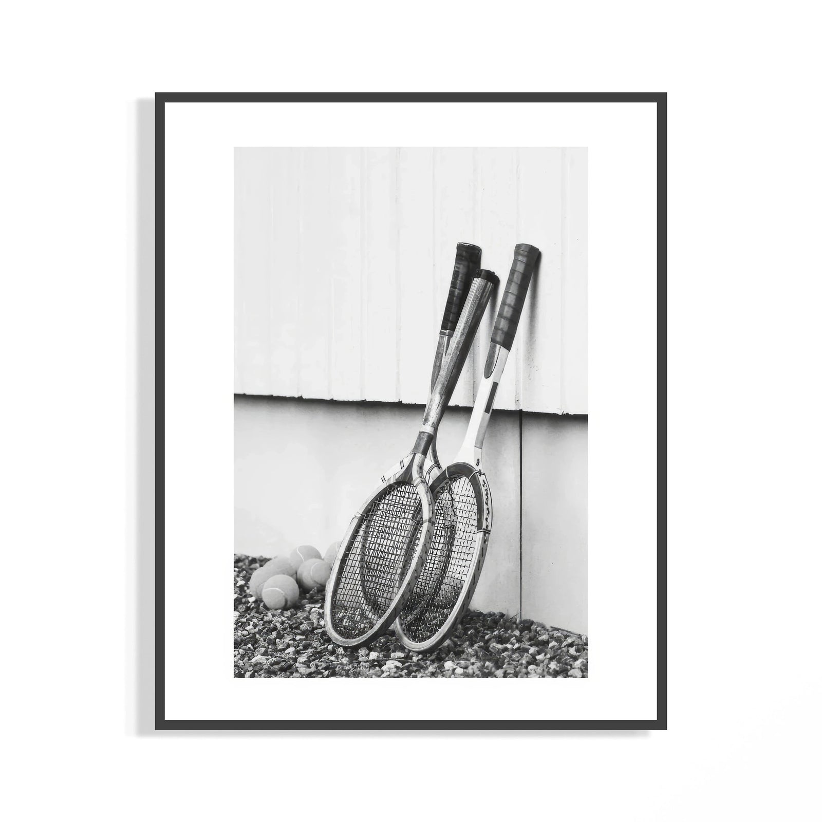 Black and white photo of vintage wooden tennis rackets leaning on a white wall over rocky ground