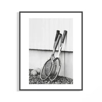 Black and white photo of vintage wooden tennis rackets leaning on a white wall over rocky ground