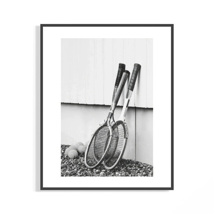 Black and white photo of vintage wooden tennis rackets leaning on a white wall over rocky ground