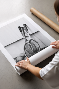 Hands unrolling a black and white wooden tennis rackets print on concrete with a shipping tube nearby