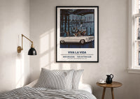 White convertible car print in a black frame above a bed with striped linens, a brass wall lamp and a sunlit window