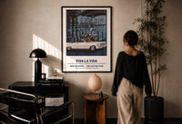 White convertible car print in a black frame on a sunlit plaster wall, a woman looking on beside a mushroom lamp