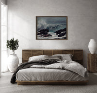 Crashing waves on rocky coast print in a dark wood frame above a rattan bed on a plaster wall