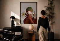 White Flowers Over the Face print in a black frame on a sunlit plaster wall, a woman looking on beside a mushroom lamp