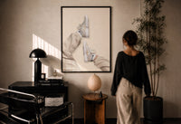 White Sneakers with Cream Trousers print in a black frame on a sunlit plaster wall, a woman looking on beside a mushroom lamp