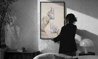 White Sneakers with Cream Trousers print in a slim black frame being hung on a sunlit wall in a monochrome room