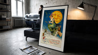 Witch riding broomstick past moon print in a slim black frame leaning on a dark sofa in a concrete print studio