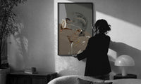 Woman in Heart Hoops and Sunglasses print in a slim black frame being hung on a sunlit wall in a monochrome room