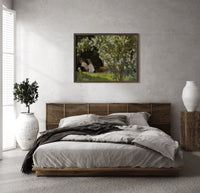 Woman reading in garden chair print in a dark wood frame above a rattan bed on a plaster wall