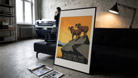 Two bighorn sheep on rocky cliff print in a slim black frame leaning on a dark sofa in a concrete print studio