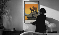 Two bighorn sheep on rocky cliff print in a slim black frame being hung on a sunlit wall in a monochrome room