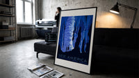 Cave stalactites with two tourists print in a slim black frame leaning on a dark sofa in a concrete print studio